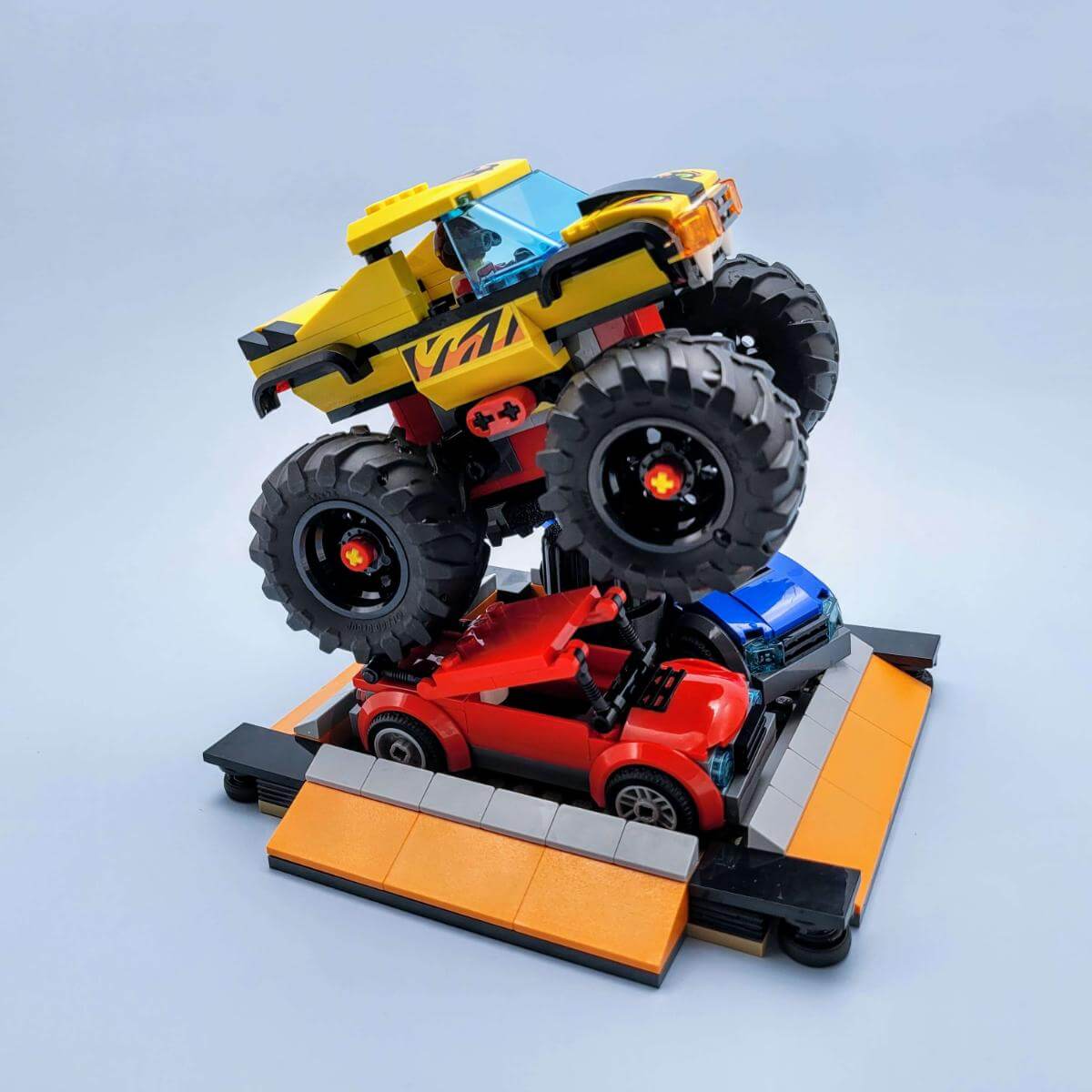 LEGO monster truck crushing a red car in a dynamic stunt scene using a black flat display base. Demonstrates how to set up action-packed displays for vehicle builds and the flexibility of mixing builds and terrain with the stand system.