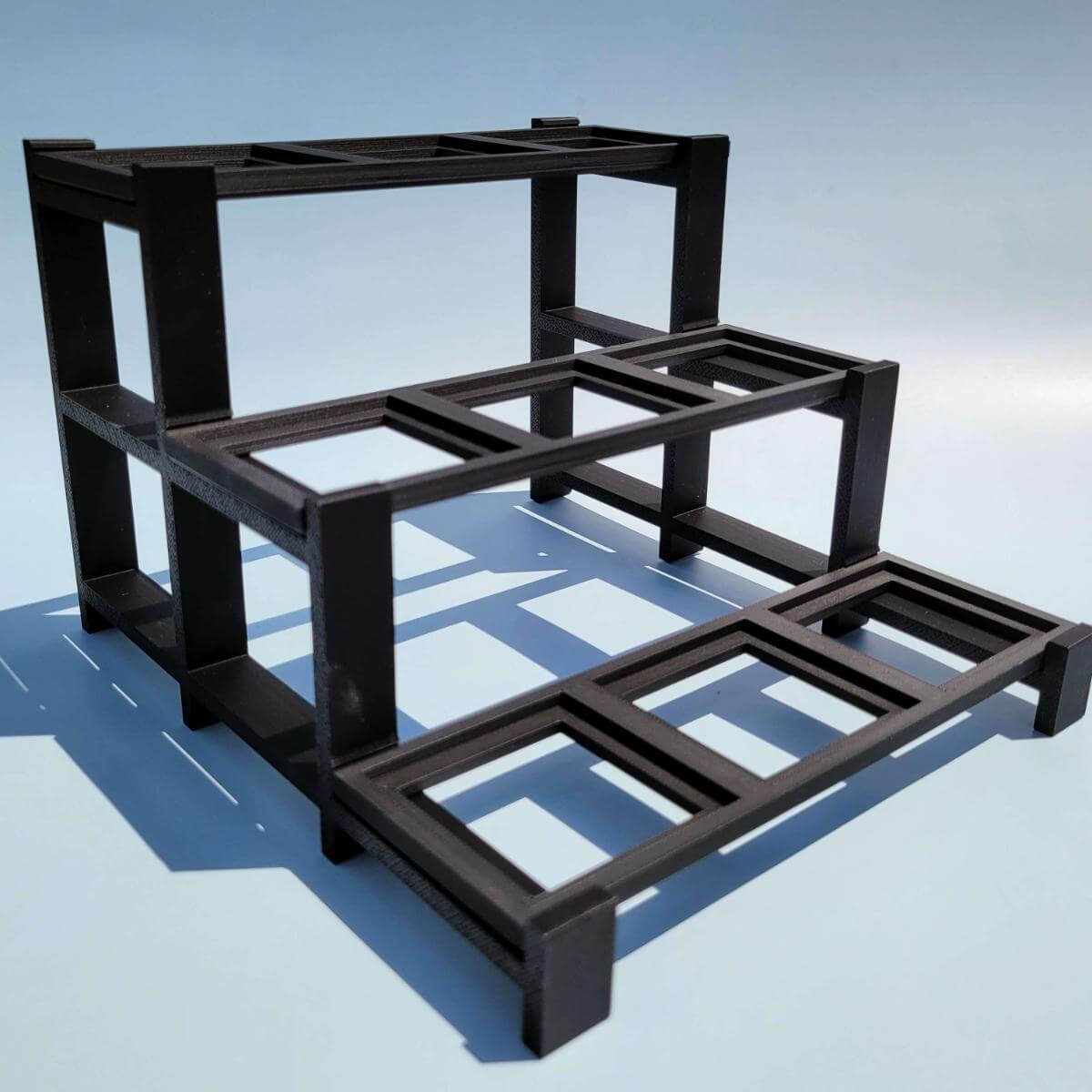 Empty three tier black LEGO compatible display stand shown from a high side angle. The stand features clean geometry with nine evenly spaced slots for displaying BrickHeadz or similar collectible figures. Perfect for minimalist display setups. Lightweight, sturdy plastic construction designed for easy assembly. A great gift for LEGO fans.
