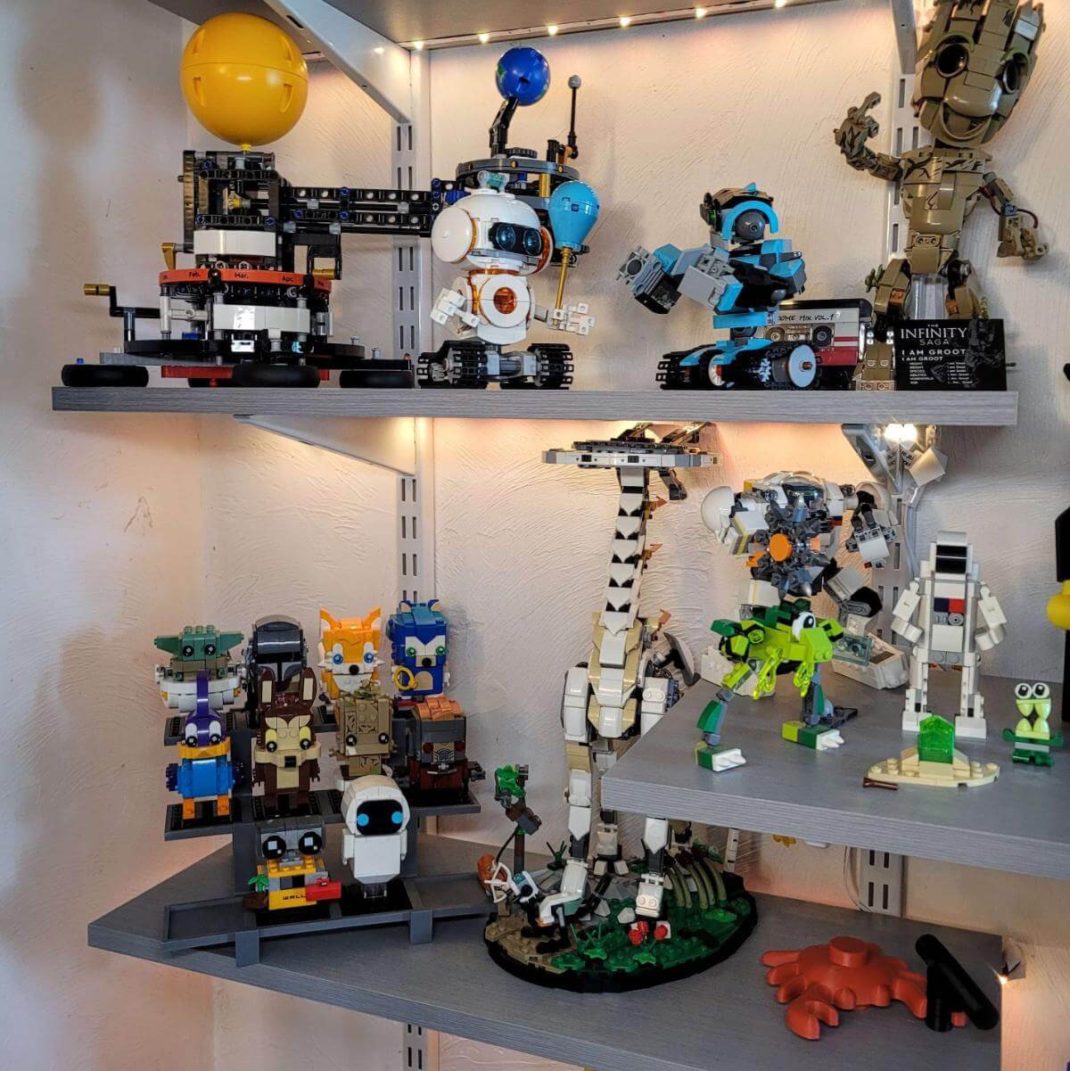 Three tier LEGO compatible display stand shown on a modern gray shelf alongside various LEGO sets, including a giraffe, robots, and Groot. The stand holds nine colorful BrickHeadz in a compact, staggered layout. Ideal for collectors looking to showcase figures in a clean and organized way. The photo demonstrates how the stand fits into a LEGO display setup. Display stand only; figures not included.