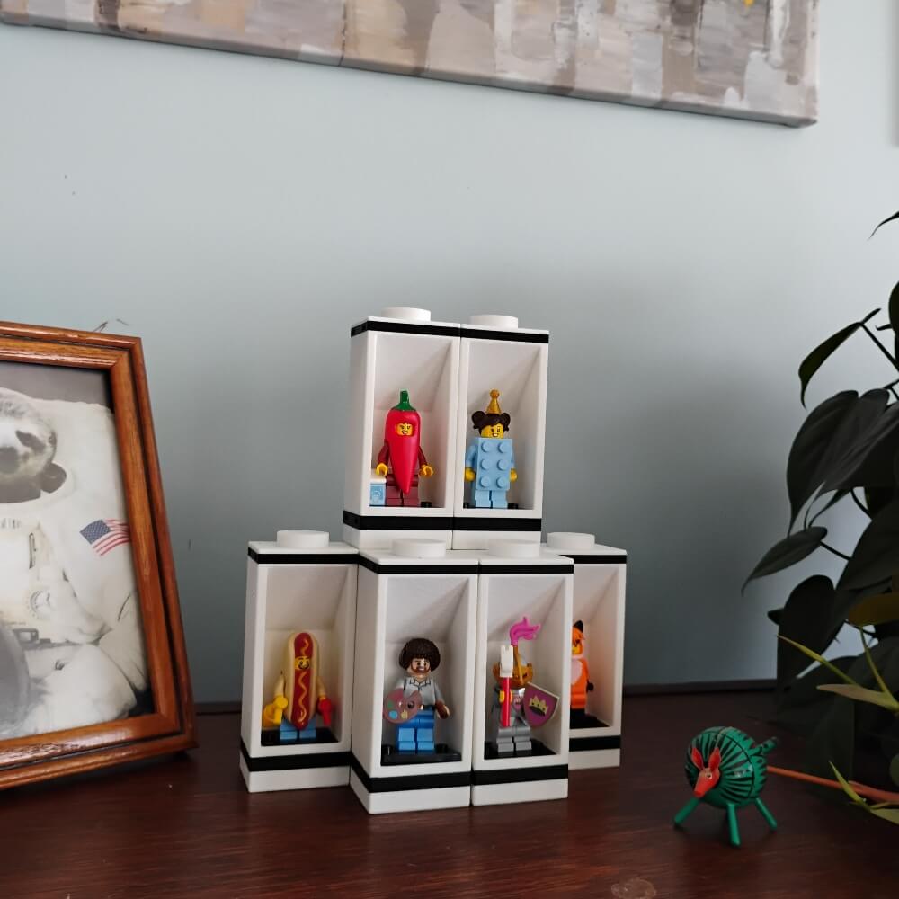 Modular LEGO® minifigure display cases arranged in a pyramid on a wooden shelf beside framed art and a plant. Each display includes a genuine LEGO® 3×4 plate to hold minifigs securely. Units stack with connector pins for stability but do not interlock, preventing damage. Perfect for AFOLs, LEGO® collectors, or as décor for desks and shelves. Clean black and white design highlights collectible figures, with custom colors available upon request.
