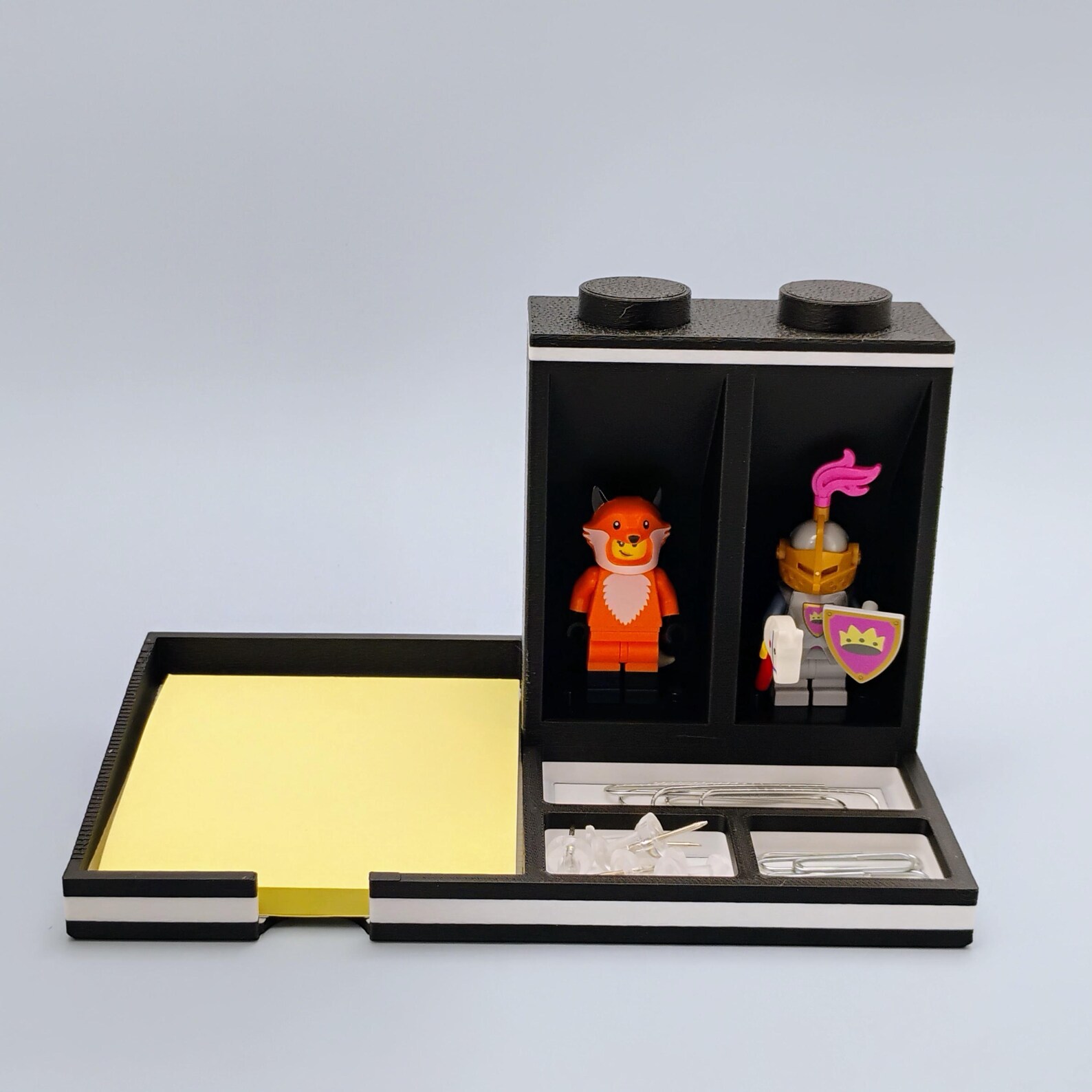 Black LEGO minifigure desk organizer with Post-it note holder. Shows the darker color option with two front-facing minifigure display areas, pen and tool storage, and a built-in sticky note tray. A clean, modern desk accessory for offices and workstations.