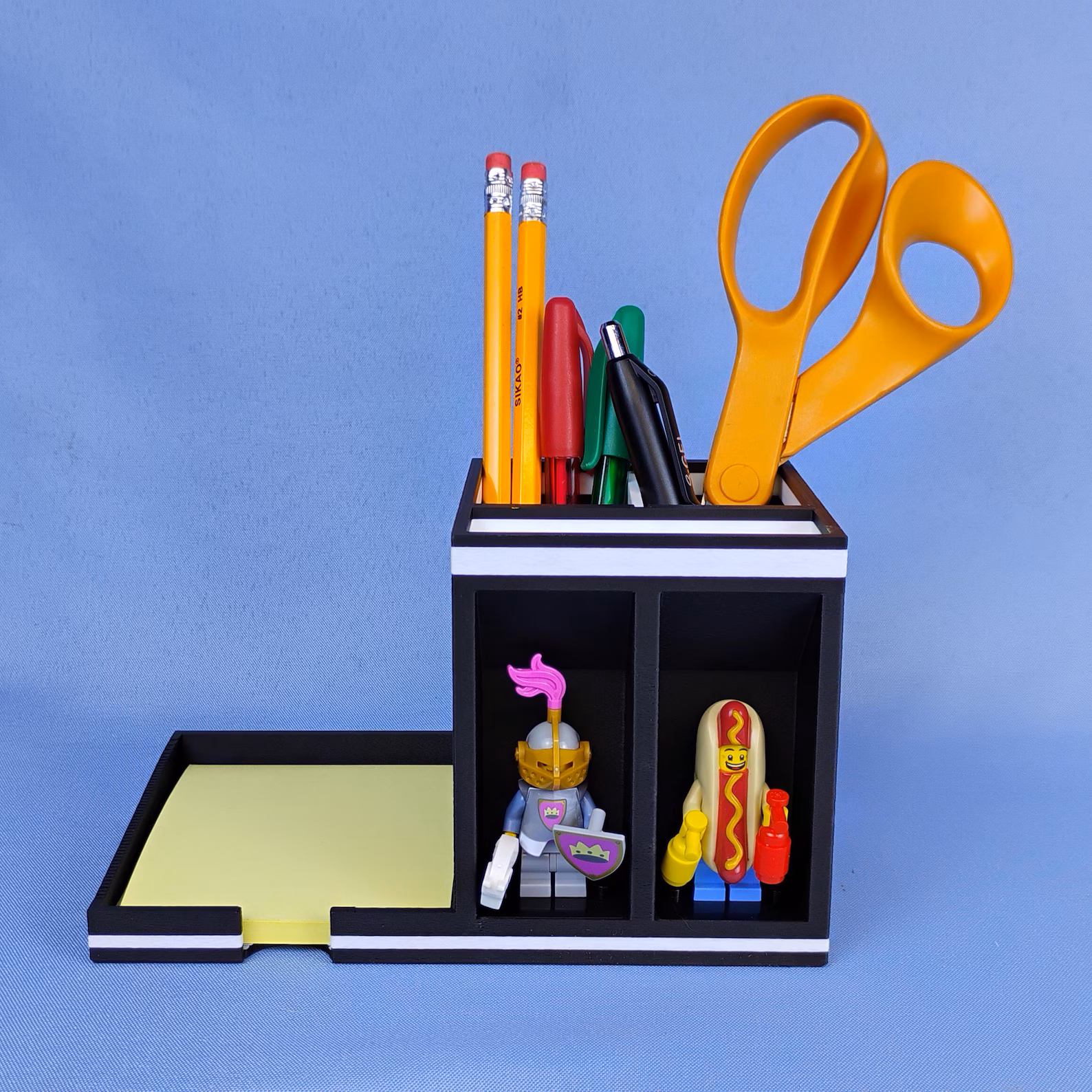 Post-it note desk organizer with pens, scissors, and sticky notes in use, featuring built-in minifigure display compartments. Demonstrates how the organizer keeps essential desk items within easy reach.