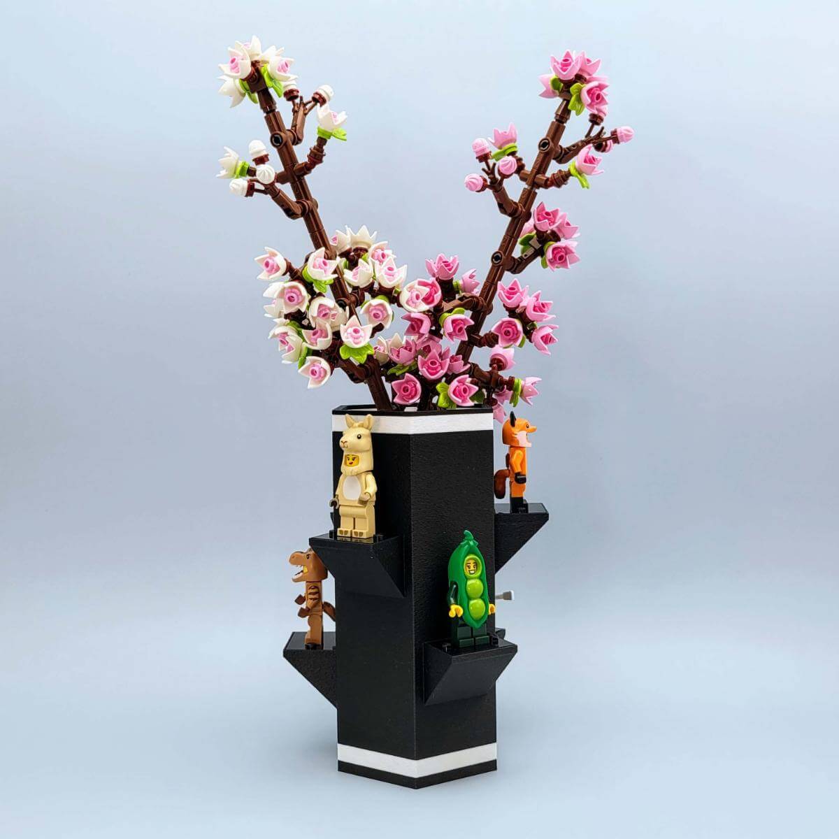 Hero image of a black LEGO compatible vase displaying vibrant cherry blossom LEGO flowers and featuring six LEGO minifigures posed on display platforms around the sides. Perfect gift for adult fans of LEGO and creative home decor enthusiasts. Combines functional display with playful LEGO integration.
