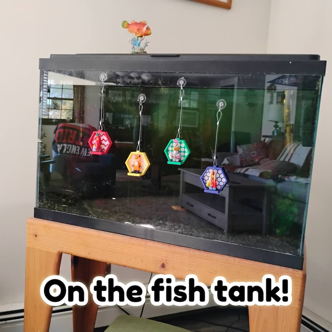Hanging LEGO® minifigure displays suspended in front of an aquarium on clear suction hooks. Bright frames pop against fish tank glass.