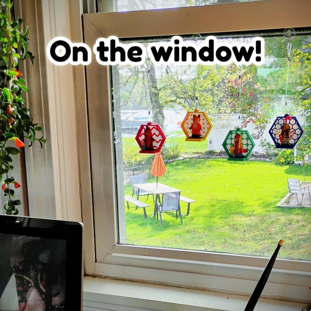 Hanging LEGO® minifigure displays on a sunny window using clear suction hooks. Colorful hexagon frames at varied heights create a bright window gallery and save shelf space.